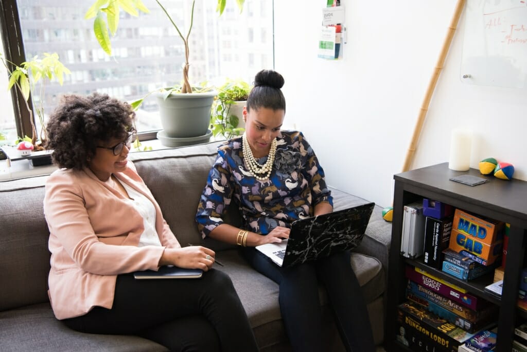 Two women are sitting on a couch at the office, pair designing on a laptop.